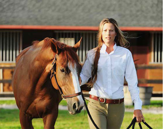 A confident equestrian woman walking beside a chestnut horse with a white blaze, holding its lead rope. The woman wears a crisp white shirt and tan riding breeches, her long hair flowing in the breeze. In the background, a stable with open stall doors and a fenced paddock adds to the serene rural setting.