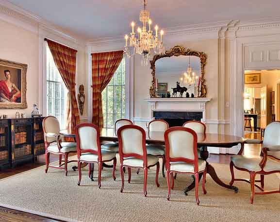 An elegant formal dining room featuring a long wooden table surrounded by upholstered chairs with red frames. A sparkling crystal chandelier hangs overhead, illuminating the space. The room showcases classic architectural details, including tall windows with plaid curtains, decorative molding, and a marble fireplace with an ornate gold-framed mirror above. A sideboard with built-in bookshelves adds charm and sophistication.