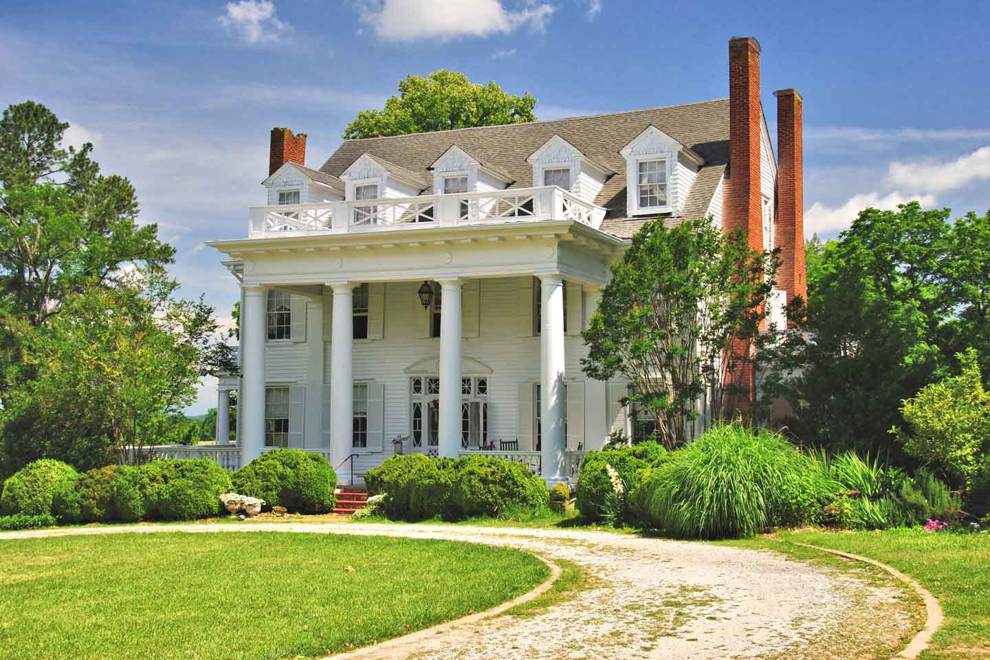 A stately historic home with classic white columns and a grand facade, featuring dormer windows, red brick chimneys, and lush landscaping in the foreground. The house exudes timeless elegance and charm under a clear blue sky. 